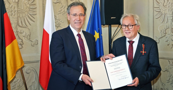 Abasador Niemiec i prof. Żyliński stoją z dyplomem orderu na tle flag Polski, Niemiec i Unii Europejskiej