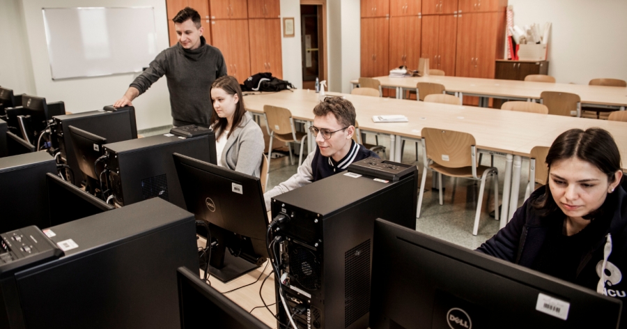 W sali komputerowej kilka osób siedzi przy stanowiskach z monitorami, pracując przy komputerach, podczas gdy jedna osoba stoi obok i wskazuje coś na ekranie, a w tle widać rzędy stołów, krzeseł i szafy.