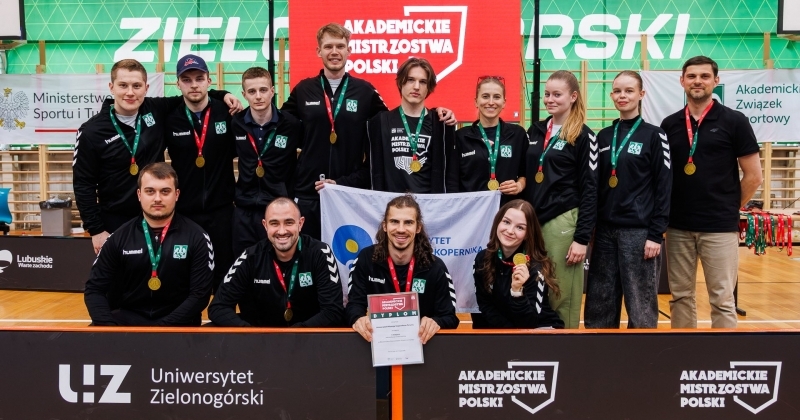 Grupa sportowców pozuje z medalami, flagą i dyplomem na tle ściany z napisem ?Uniwersytet Zielonogórski? i ekranu ?Akademickie Mistrzostwa Polski?.
