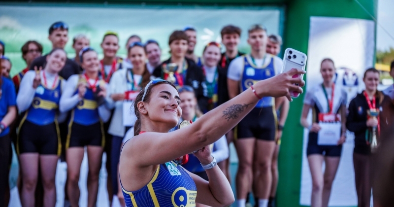 Grupa uśmiechniętych wioślarek i wioślarzy AZS UMK Toruń w strojach sportowych pozuje wspólnie do zdjęcia, trzymając w rękach zdobyte puchary i dyplomy. Jedna z wioślarek trzyma telefon, robiąc nim zdjęcie.