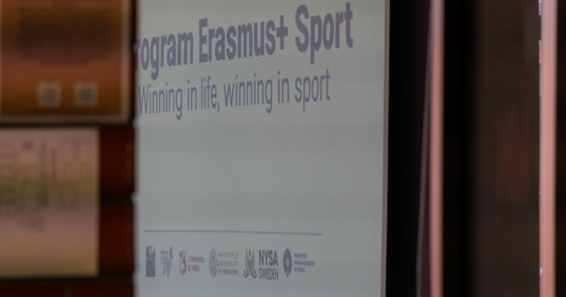 Zdjęcie zrobione z ukosa, przedstawiające ekran projekcyjny w Sali Audytoryjnej. Na ekranie widoczny jest fragment napisu ?Program Erasmus+ Sport? oraz poniżej podtytuł ?Winning in life, winning in sport?. Na dole slajdu umieszczone są logotypy instytucji partnerskich projektu. Lewa strona kadru jest częściowo zasłonięta przez rozmazany, pierwszy plan z fragmentem ściany.