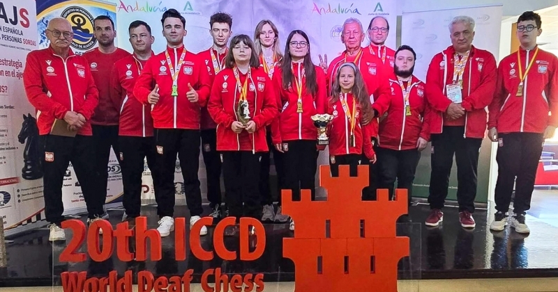Reprezentacja Polski w szachach na podium w czerwono-białych strojach narodowych z medalami i pucharami. Na pierwszym planie stoi przezroczysta tablica z czerwonym napisem ?20th ICCD World Deaf Chess Championships Roquetes de Mar ? SPAIN 2026? oraz logo w kształcie zamku na tle szachownicy.