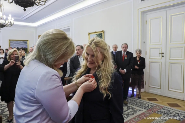 Kobieta przypina odznaczenie drugiej kobiecie podczas oficjalnej uroczystości w eleganckiej sali, w tle stoją zaproszeni goście.