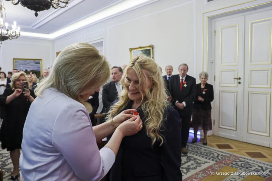 Kobieta przypina odznaczenie drugiej kobiecie podczas oficjalnej uroczystości w eleganckiej sali, w tle stoją zaproszeni goście.