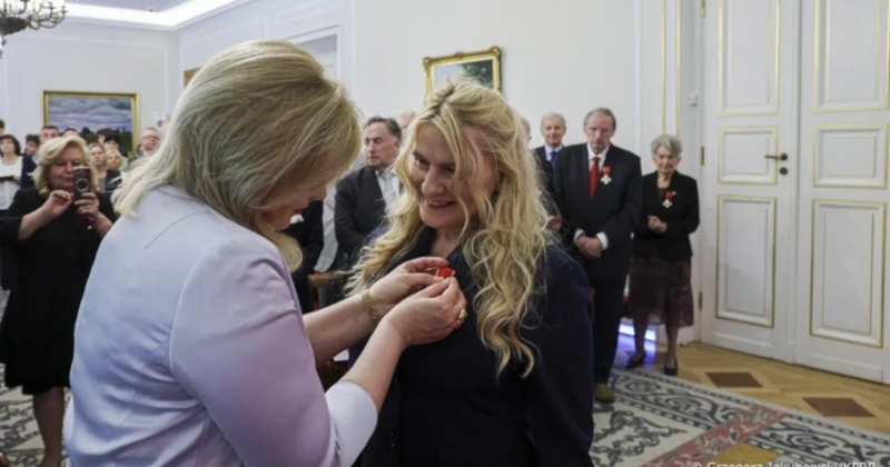 Kobieta przypina odznaczenie drugiej kobiecie podczas oficjalnej uroczystości w eleganckiej sali, w tle stoją zaproszeni goście.