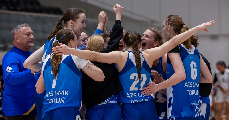 Grupa koszykarek w niebieskich strojach meczowych z napisami ?AZS UMK TORUŃ? oraz ich trener w niebieskiej bluzie stoją w ciasnym kółku na parkiecie hali sportowej, krzycząc i obejmując się z uniesionymi w górę rękami.