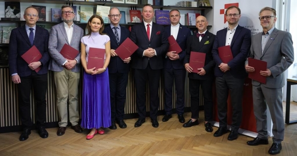 Ośmiu mężczyzn i jedna kobieta uroczyście ubrani stoją z aktami powołania do rady na tle regału z ksiażkami