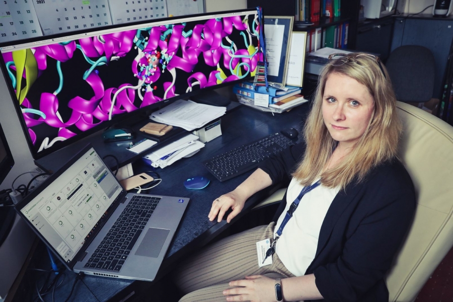 Dr. hab. Karolina Mikulska-Rumińska, NCU Prof., sitting at a desk in an office. On the desk, there is a laptop and a large monitor displaying a colorful 3D model of a protein structure.