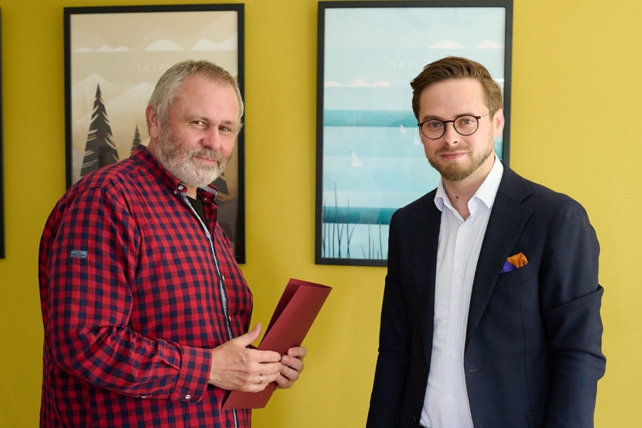 Na tle żółtej ściany z kolorowymi grafikami w ramkach pozuje do zdjęcia dwóch mężczyzn - z lewej starszy w czerwono-czarnej koszuli, z tekturową teczką w dłoniach, z prawej młodszy, w okularach, w czarnej marynarce i białej koszuli.